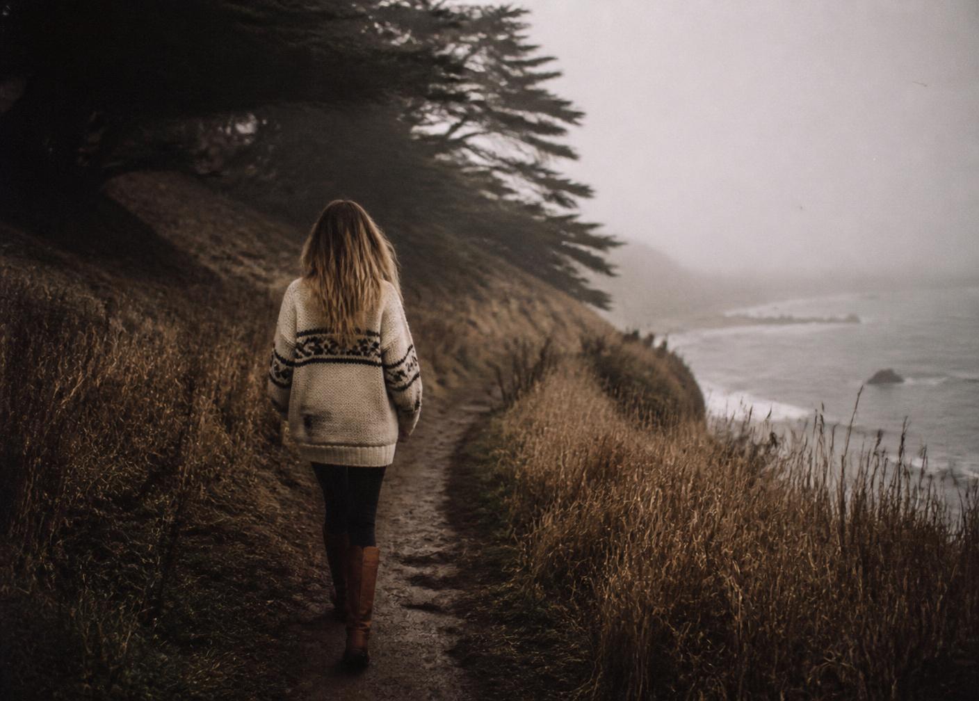 A woman in an oversized wool sweater and worn boots walking a foggy Point Reyes coastal trail in winter, seen from behind
